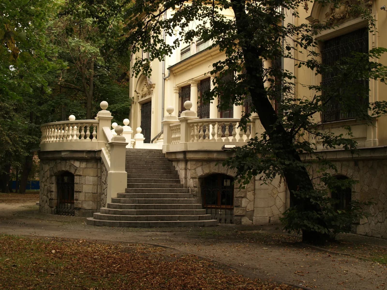 A grand stone staircase with a decorative balustrade leads up to a light-colored building with dark windows, surrounded by green trees and fallen autumn leaves.
